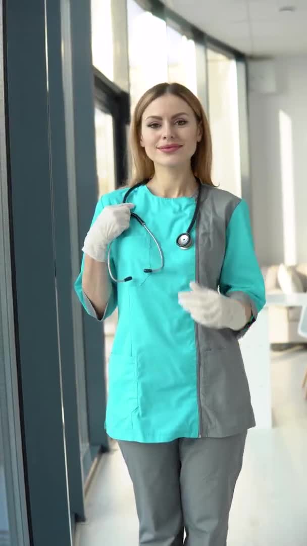 Seorang dokter wanita mengenakan mantel medis dan stetoskop melihat ke kamera. Tersenyum dokter perempuan atau perawat di kantor rumah sakit. Video vertikal — Stok Video
