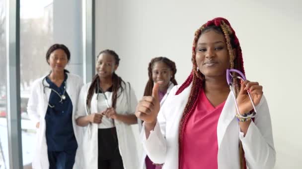 Un joven médico afroamericano con el pelo rojo se para delante y mira a la cámara en el fondo de un equipo de colegas — Vídeo de stock