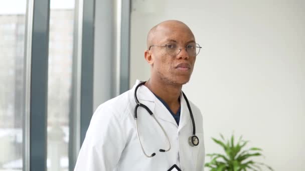 Ένας νεαρός αφροαμερικανός γιατρός στέκεται με ιατρική στολή στην κλινική και κοιτάζει την κάμερα. — Αρχείο Βίντεο