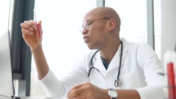 Un tânăr medic afro-american care lucrează cu sângele în tubul de testare de la birou. Dezvoltarea vaccinului covid 19 — Videoclip de stoc