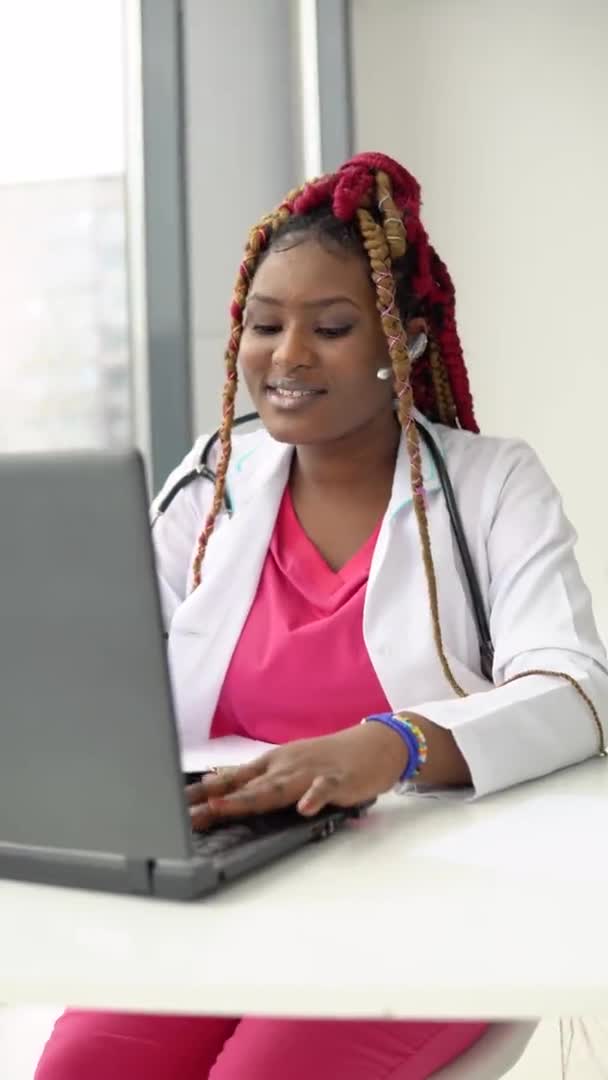 Junge afrikanisch-amerikanische Ärztin mit roten Haaren plaudert oder berät sich am Laptop. Vertikales Video — Stockvideo