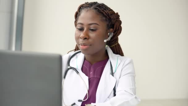 Junge afrikanisch-amerikanische Ärztin mit Headset bei Chat oder Konsultation am Laptop — Stockvideo