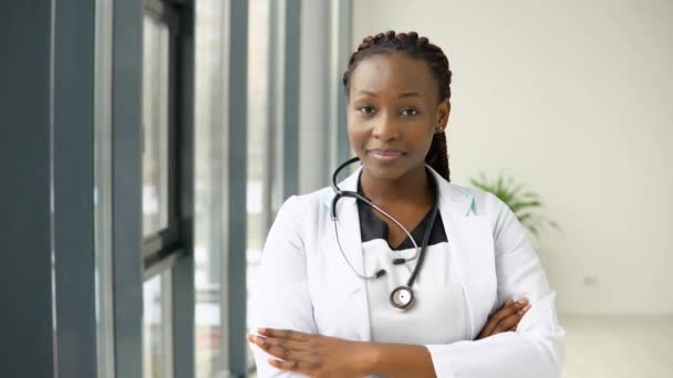 Молодая африканская женщина врач или медсестра, стоящая в медицинском костюме в клинике и смотрящая в камеру — стоковое видео