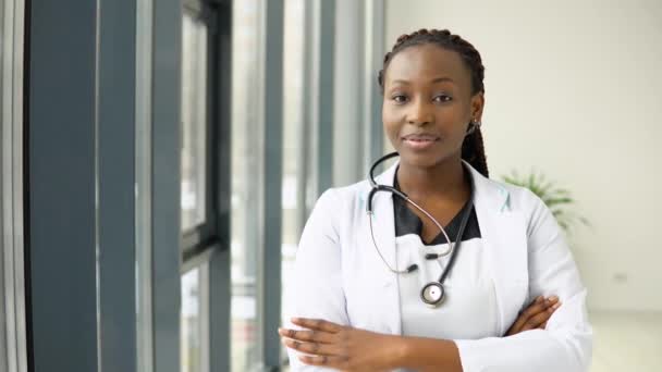 Молодая африканская американка врач или медсестра, стоящая в медицинском костюме в клинике и смотрящая в камеру — стоковое видео