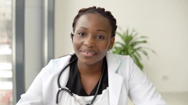 Genç Afrikalı Amerikalı kadın doktor bilgisayarda sohbet ediyor ya da danışmanlık yapıyor. Doğrudan kameraya bakıyor.