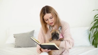 Rahat toz pembe bir kazak ve beyaz çoraplı genç ve güzel bir kadın köpeğiyle birlikte yatağa oturur ve bir kitap okur.