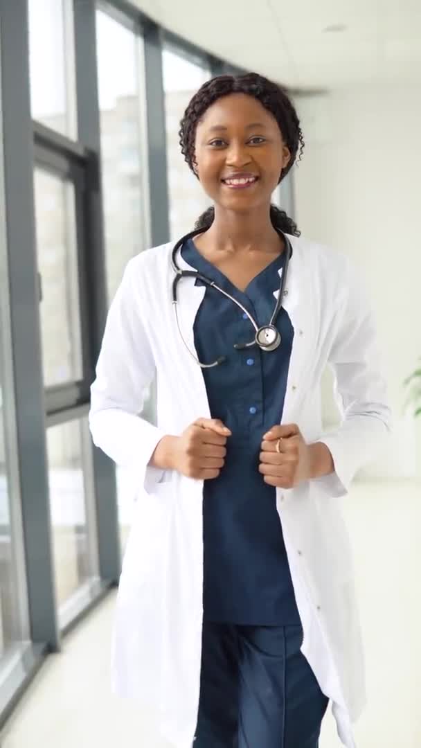 Μια νεαρή Αφροαμερικάνα γιατρός ή νοσοκόμα στέκεται με ιατρική στολή στην κλινική και κοιτάζει την κάμερα. Κάθετη βίντεο — Αρχείο Βίντεο