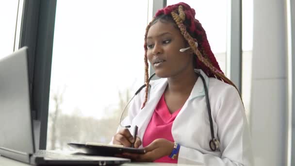 Jovem afro-americana médica com cabelo ruivo tendo bate-papo ou consulta no laptop — Vídeo de Stock