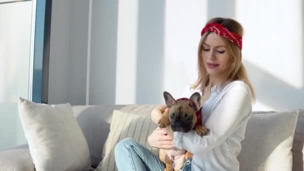 Mujer joven en una camisa blanca y jeans se sienta en el sofá y juega con su perro — Vídeo de stock