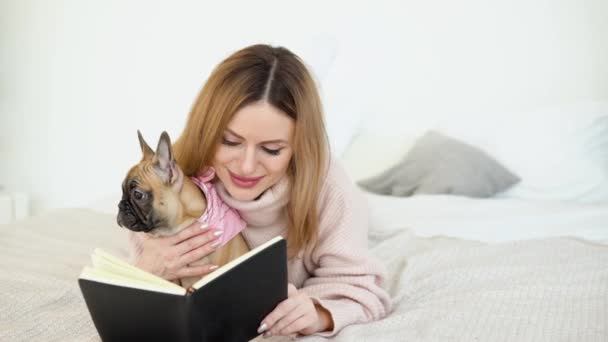 En kvinna i en mysig puderrosa tröja och vita strumpor liggande på sängen med sin hund och läser en bok — Stockvideo