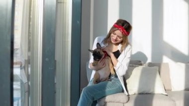 Rahat bir evde, kollarında bir buldog köpeğiyle rahat rahat oturan genç bir kadın, parlak güneşin altında bir kanepede oturuyor.