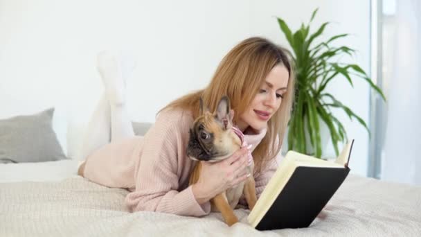 Een jonge vrouw in een knusse poederroze trui en witte kousen liggend op het bed met haar hond en leest een boek — Stockvideo