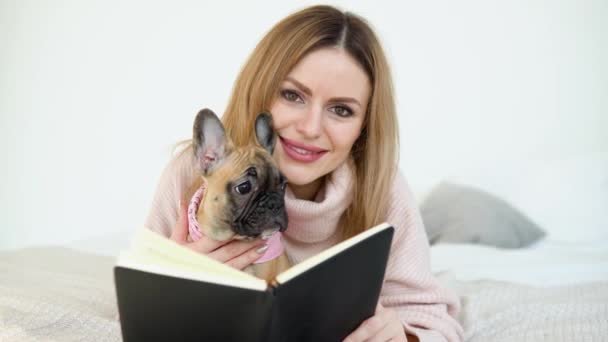 Een jonge vrouw in een knusse poederroze trui en witte kousen liggend op het bed met haar hond en een boek in haar handen kijkt naar de camera — Stockvideo