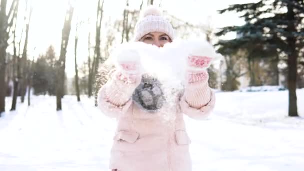 女人吹雪。穿着柔软粉色羽绒服的年轻女子在雪地的背景下在摄像机前吹雪 — 图库视频影像