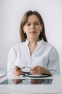 Beyaz ceketli ve steteskoplu beyaz kadın doktor kameraya bakıyor. Hastane ofisinde poz veren gülümseyen kadın doktor.