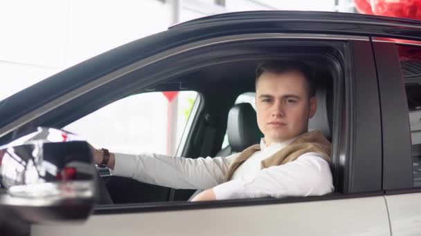 Πορτρέτο ενός νεαρού άνδρα με αυτοπεποίθηση κάθεται σε ένα νέο αυτοκίνητο σε μια αντιπροσωπεία αυτοκινήτων — Αρχείο Βίντεο