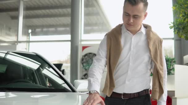 Ο τύπος αγγίζει την κουκούλα ενός γυαλιστερού λευκού αυτοκινήτου. Νεαρός άνδρας κοιτάζει προσεκτικά το αυτοκίνητο στην αντιπροσωπεία αυτοκινήτων — Αρχείο Βίντεο
