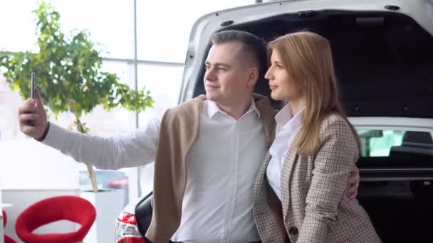 Pareja feliz tomando una selfie en un concesionario de coches con un coche nuevo en el fondo — Vídeo de stock