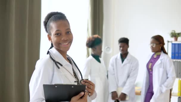 Beyaz önlüklü ve steteskoplu Afrikalı Amerikalı kadın doktor kameraya bakıyor ve klinikte gülümsüyor. Arkaplandaki doktorlar — Stok video