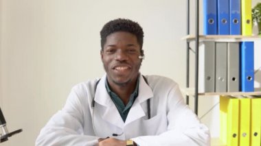 Genç Afro-Amerikan erkek doktor bilgisayarda sohbet ediyor ya da danışmanlık yapıyor doğrudan kameraya bakıyor. Kendine güvenen genç doktor seminer düzenliyor, giyiniyor, ders veriyor.