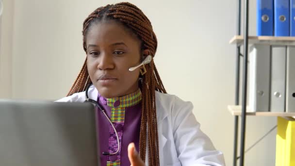 Jovem afro-americana médica com fone de ouvido tendo bate-papo ou consulta no laptop — Vídeo de Stock