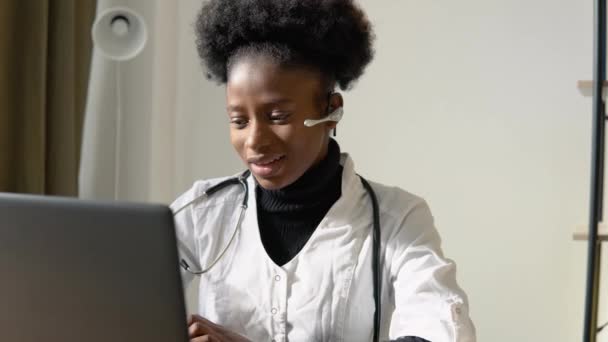 Junge afrikanisch-amerikanische Ärztin mit Headset beim Chatten oder Konsultieren auf dem Laptop. Ärztin trägt Kopfhörer und hält medizinische Online-Konferenz ab — Stockvideo