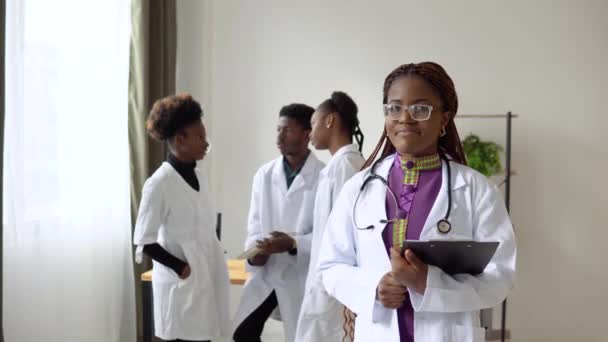 Una giovane dottoressa afroamericana si trova di fronte e guarda la telecamera sullo sfondo di un team di colleghi — Video Stock