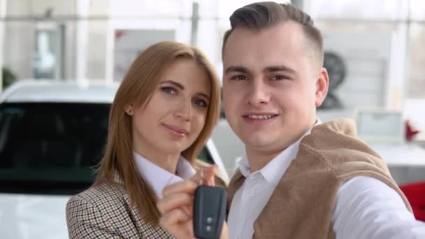 Happy couple taking a selfie at a car dealership with a new car in the background. Man and woman showing keys of his new car in dealership — Stock Video