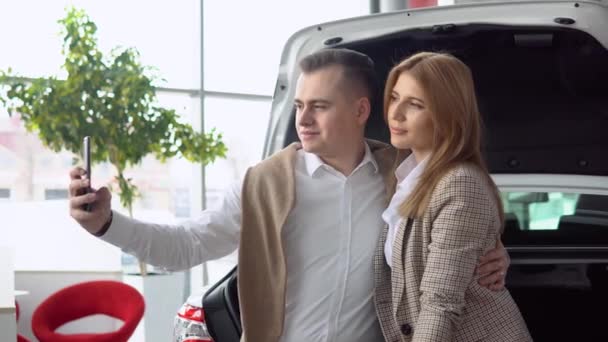 Coppia felice scattare un selfie in una concessionaria di auto con una nuova auto sullo sfondo — Video Stock