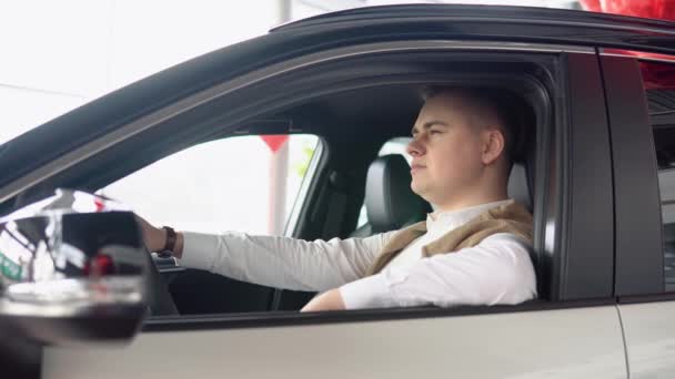Porträtt av en ung självsäker man som sitter i en ny bil hos en bilhandlare. Snygg man inspekterar en ny bil hos en bilhandlare — Stockvideo