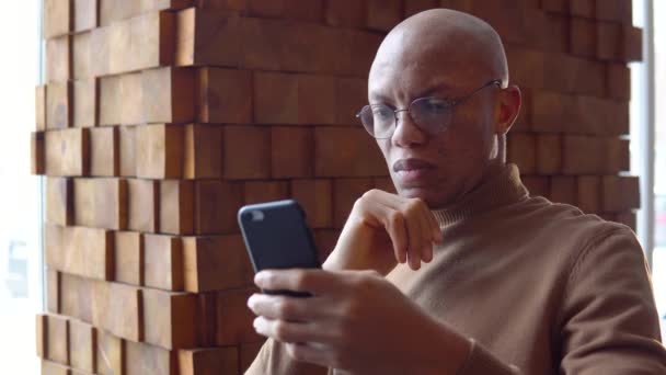 Серьезный молодой африканский мужчина, работающий в кафе со смартфоном — стоковое видео