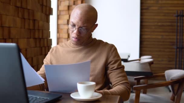 Człowiek z poważnym wyrazem twarzy pracuje z laptopem i dokumentami w kawiarni. Estetyka karmelowa. Pandemia — Wideo stockowe