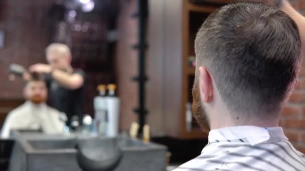 Um barbeiro em uma barbearia faz penteado para um cliente. Vista no espelho — Vídeo de Stock