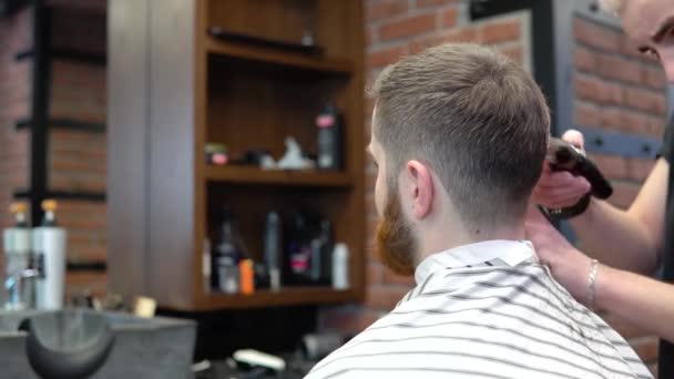 Un joven peluquero hace un corte de pelo de un cliente con barba roja — Vídeos de Stock