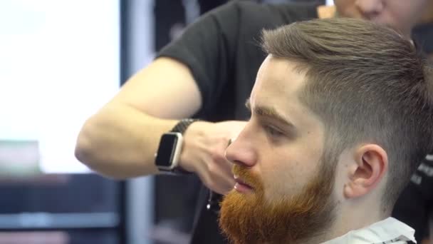 Um jovem com barba está sentado em uma cadeira de cabeleireiro durante um corte de cabelo. Vista lateral — Vídeo de Stock