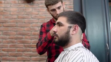 Ekose kırmızı ve siyah gömlekli genç bir kuaför berber dükkanında müşterilerin kafasının arkasını kesiyor.