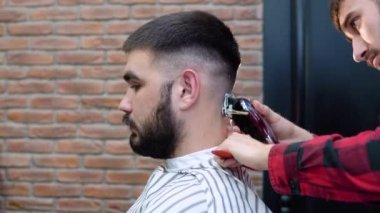 Berber, müşterinin kafasının arkasında saç kesme makinesi kesiyor. Koyu saçlı, genç, beyaz adamlar. Görüntüyü kapat