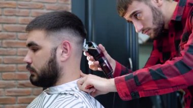 Berber, müşterinin kafasının arkasında saç kesme makinesi kesiyor. Koyu saçlı, genç, beyaz adamlar. Görüntüyü kapat