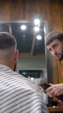 Genç berber müşteriyi çok dikkatli bir şekilde budayıcı ile keser. Vip Berber Dükkanı. Dikey video.