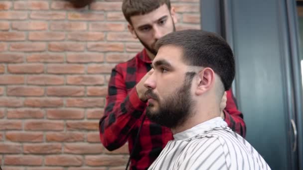 Joven peluquero elegante en una camisa a cuadros rojo y negro hace un corte de pelo en la parte posterior de la cabeza de los clientes en una barbería — Vídeos de Stock