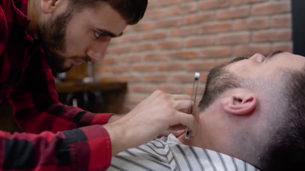 Penata rambut dengan lembut mencukur jenggot klien dengan pisau cukur berbahaya. Pria Kaukasia. Barbershop — Stok Video