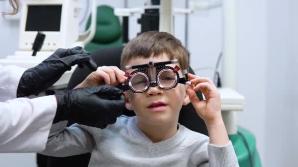 De oogarts maakt aanpassingen aan het proefframe om lenzen te selecteren voor de blanke jongen — Stockvideo