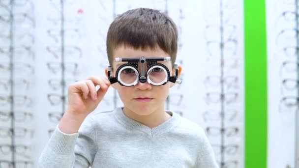 Petit garçon se tient sur un fond de vitrine avec montures pour lunettes avec le dispositif de sélection des lentilles de contact — Video