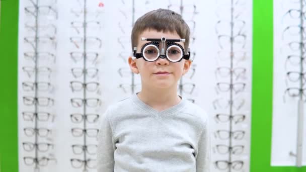 Mały chłopiec stoi na tle okna wystawowego z ramkami na okulary z urządzeniem do doboru soczewek kontaktowych — Wideo stockowe