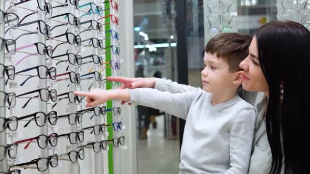 Молодая белая женщина с ребенком, мать и сын выбирают очки в магазине оптики — стоковое видео