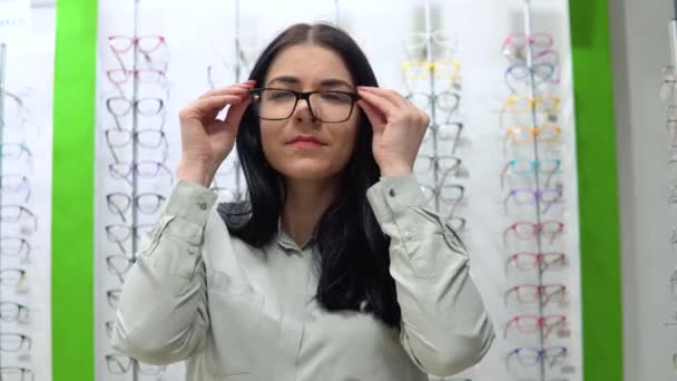Νεαρή Καυκάσια γυναίκα δοκιμάζει γυαλιά στο φόντο μιας βιτρίνας καταστήματος με πλαίσια θεάματος — Αρχείο Βίντεο