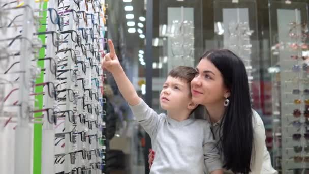 Mujer joven con bebé, madre e hijo eligiendo gafas en la tienda de óptica. Óptica, tienda óptica — Vídeos de Stock