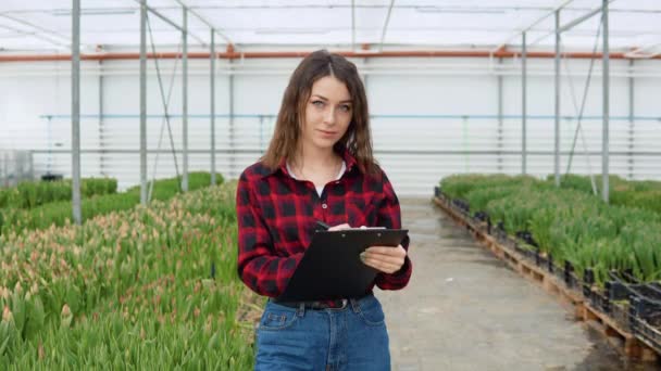 Цветочный молодой кавказской девушки в красно-черной рубашке и джинсах стоит в оранжерее с тюльпанами с табличкой в руках — стоковое видео