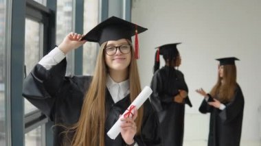 Bir elinde diploma, diğer elinde farklı ırktan iki öğrencinin arka planında şapka tutan neşeli bir mezun. Orta sınıf kadınlar için yüksek öğrenim mevcuttur