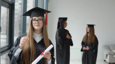 Üstün cüppeli bir öğrenci kameranın önünde duruyor ve arka planda iki öğrenci iletişim kuruyor. Üniversite eğitiminin başarısı. Bilimin Efendisi